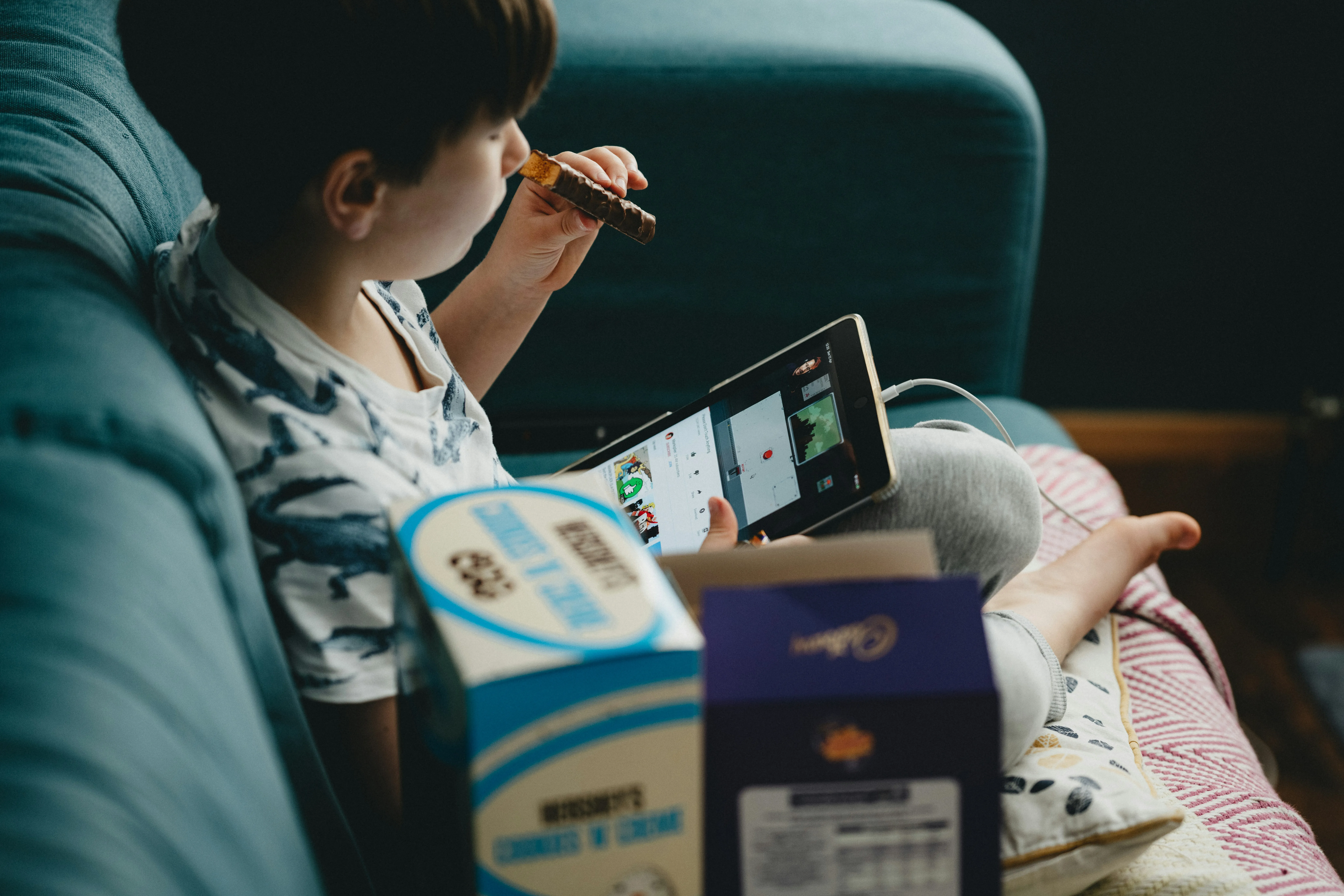 A child curled on a teal couch watches a tablet propped against their knees, snack in hand, boxes of candy in the foreground.