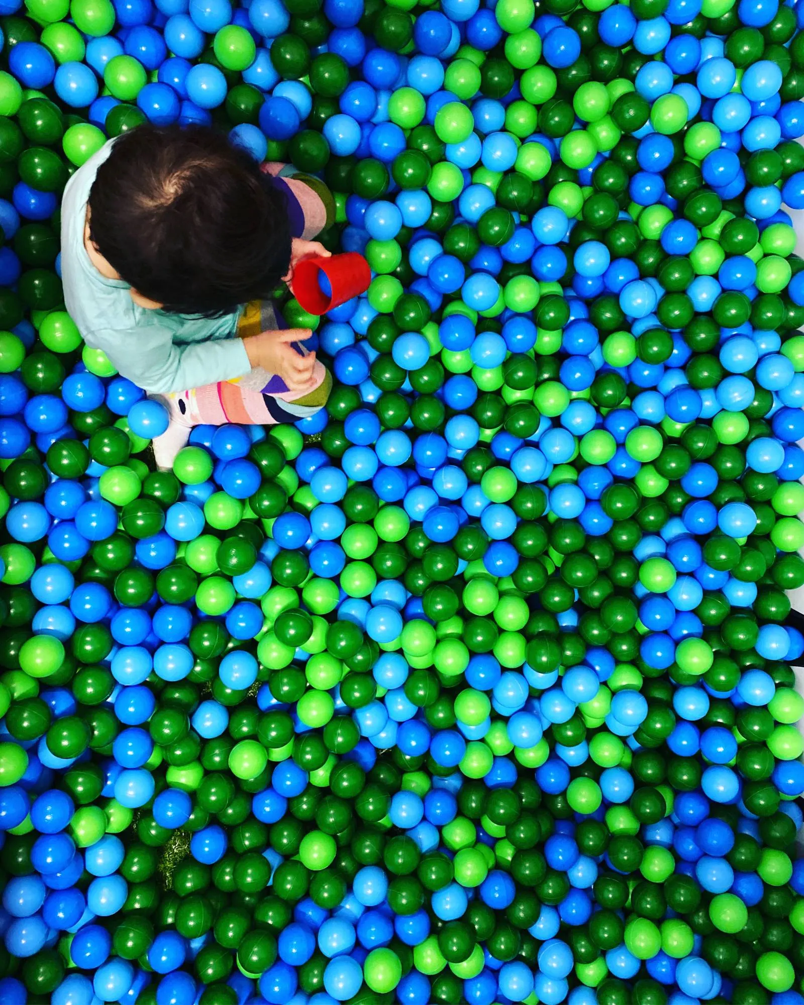 A young child seen from above, sitting in a ball pit of blue and green balls, holding a red cup.