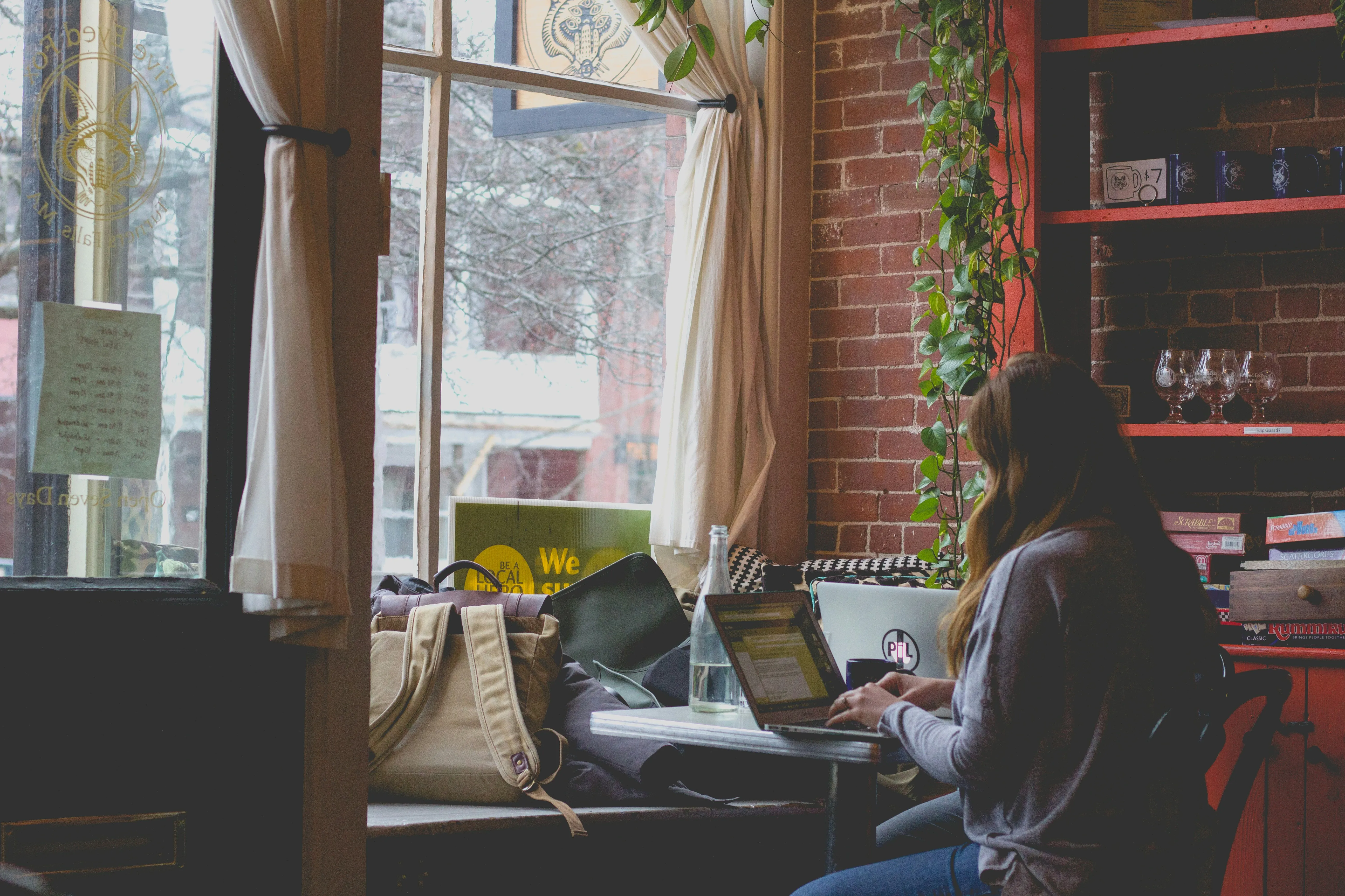 A woman reading on a laptop at a cafe window, beside a shelf of board games and a trailing houseplant.