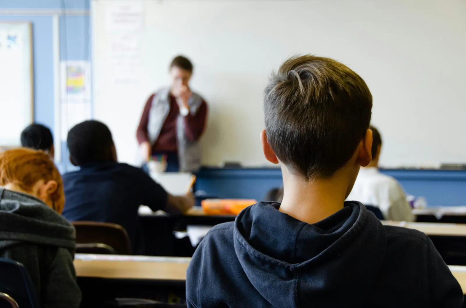 Seen from the back of a classroom, students at desks facing a teacher standing at the whiteboard.