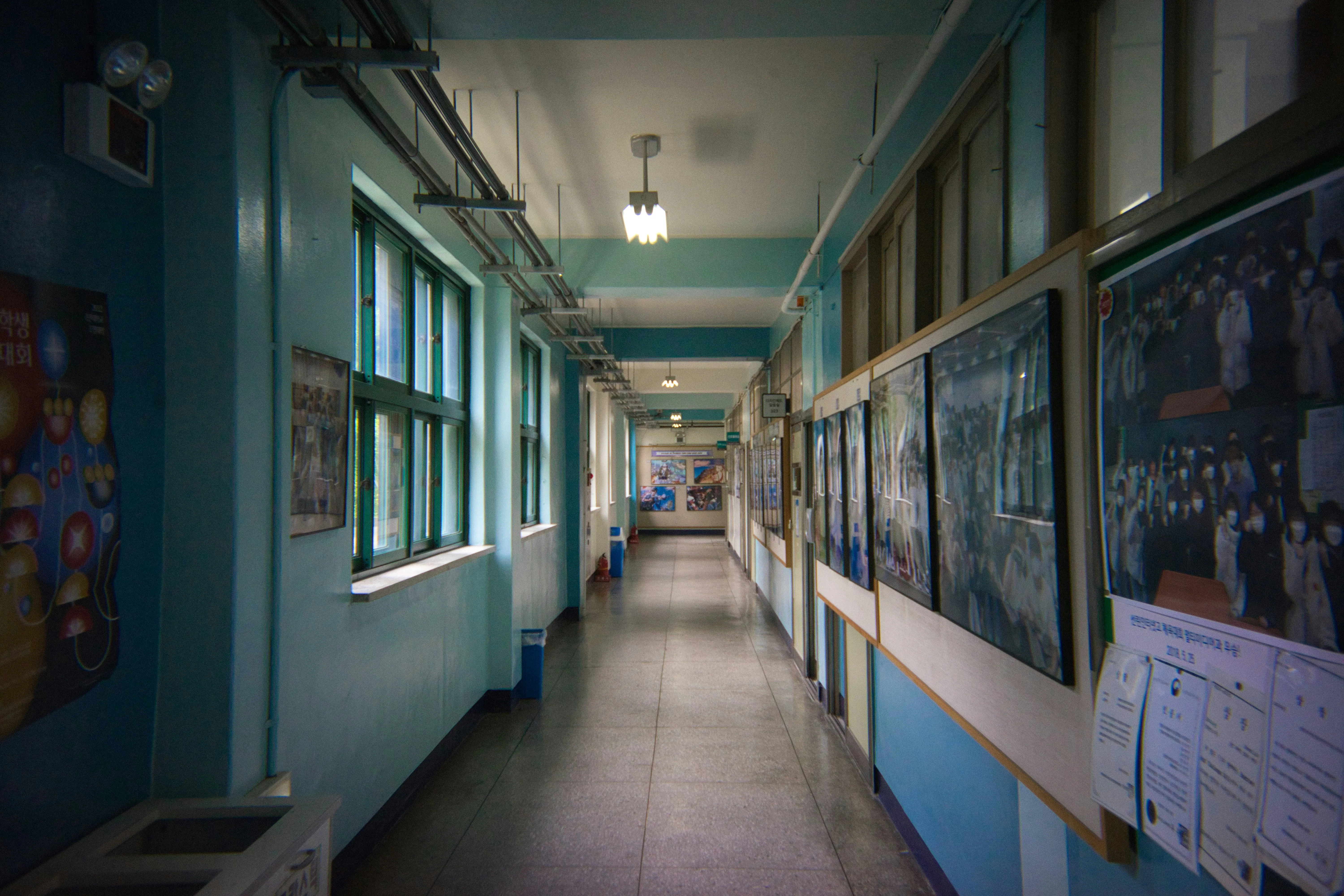An empty school hallway with pale blue walls, green-framed windows down one side, and bulletin boards of class photos and notices down the other.