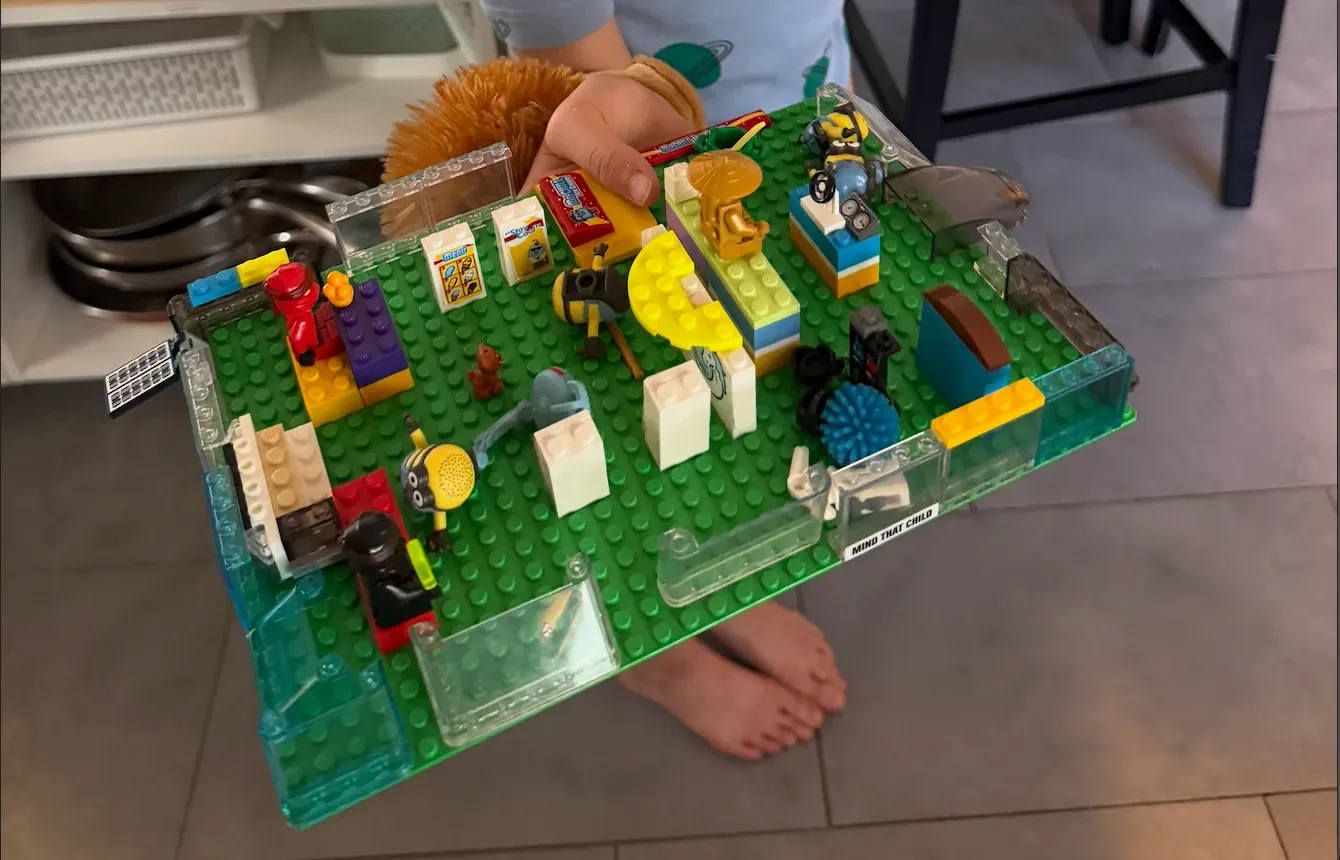 A child holds up a small world built on a green Lego baseplate, dense with figures and furniture.