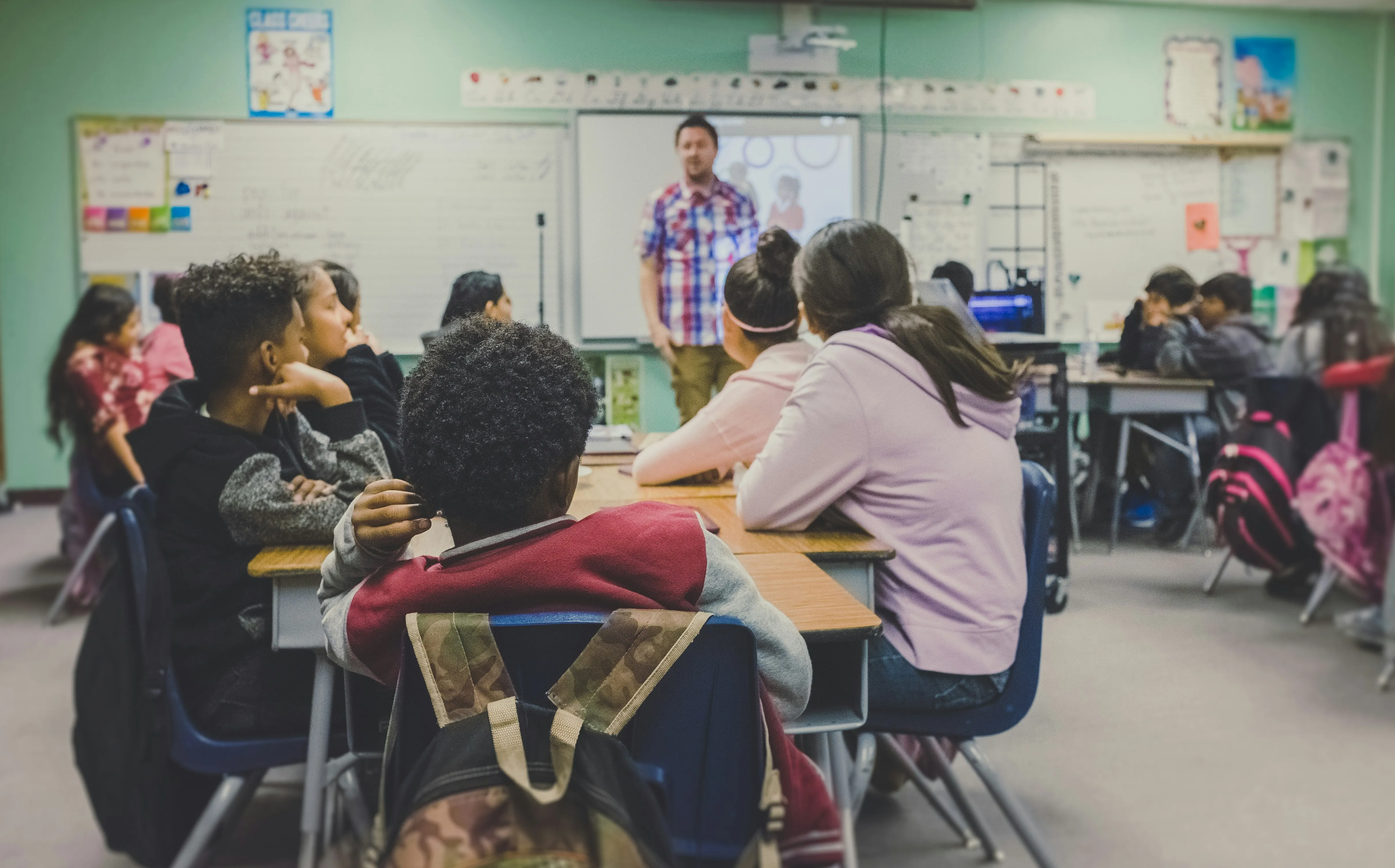 Seen from the back of a classroom, students at desks facing a teacher standing at the whiteboard.