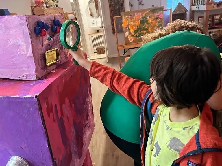 A child holds a magnifying glass up to the face of a robot built from cardboard boxes, painted purple and pink, with rows of blue and red knobs and a gold dial.