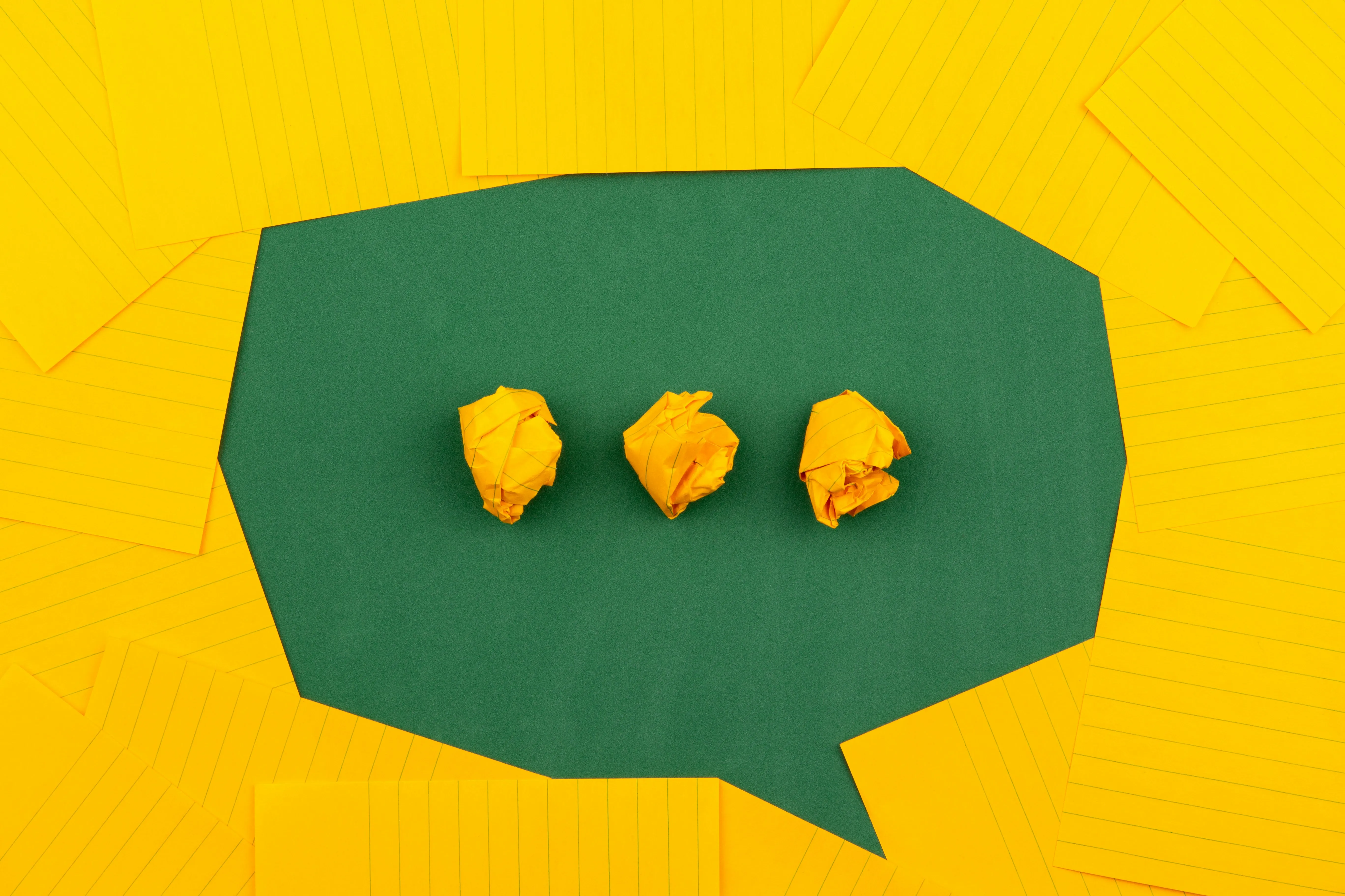 A green paper speech bubble on a bed of yellow lined notepaper, holding three crumpled yellow paper balls.